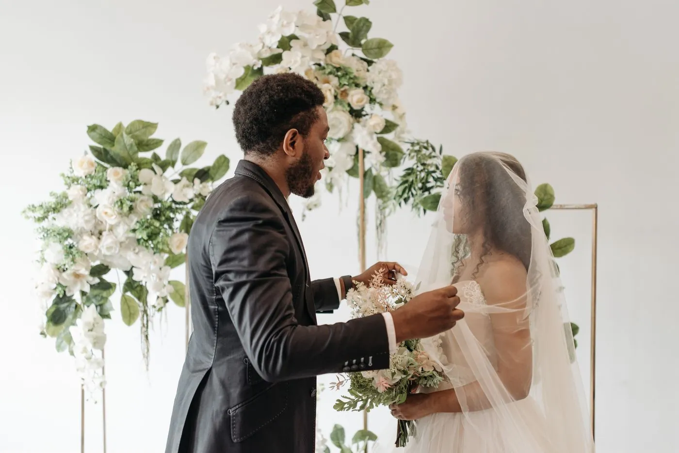 A groom lifting the bride's veil at a floral ceremony arch